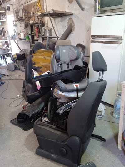 The image depicts a cluttered workshop environment in Sukleya, Moldova. The foreground is dominated by disassembled car seats, including a child car seat with the brand "Alto" visible. One car seat is partially dismantled, revealing its internal structure, and another has a detached headrest. Various car parts, such as bumpers and panels, are scattered around, some appearing to be made of plastic and metal. Yellow and gray automotive components are piled high. In the background, a white refrigerator with a vintage television on top stands against a concrete wall. The workshop walls are adorned with an assortment of tools, including wrenches, pliers, and hammers, neatly arranged on a board. Other items visible include a roll of paper towels, paint cans, and various wires and cables on the floor. The lighting suggests it is daytime. There are no people visible in the scene.