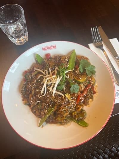 An overhead shot captures a dark stir-fry dish served on a white plate with a red rim, branded "BALTHAZAR FOOD & DRINKS," positioned on a dark table surface. The dish consists of dark-coated rice or noodles, mixed with green snap peas, sliced zucchini or cucumber, red bell peppers, and other vegetables, garnished with white sesame seeds, a cluster of white bean sprouts, and fresh green cilantro sprigs. To the left of the plate, a clear, textured glass tumbler filled with water and ice cubes is visible. To the right of the plate, a silver fork and knife rest on a folded white napkin. A dark, woven placemat or table texture is partially visible in the bottom right corner. The setting is indoors, specifically at Balthazar Food & Drinks, located at Grote Markt 32, 8500 Kortrijk, Belgium. No individuals are depicted, and no direct actions or interactions are occurring.