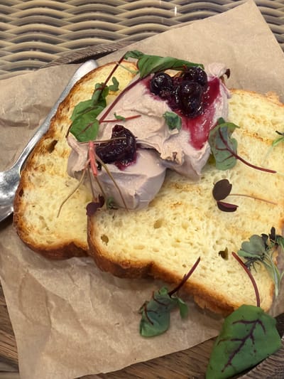 This is a close-up, overhead shot of two slices of toasted bread topped with a spread of pale pink pâté. The pâté is garnished with dark berries and a red sauce, as well as microgreens with red veins. A silver spoon is visible on the left side of the frame, resting on brown parchment paper. The food is placed on a woven surface. The scene is indoors, suggesting it might be a restaurant or cafe. The image does not provide any information to determine its location in Arkhangel'skoye, Russia.