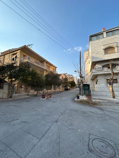 This image depicts a quiet residential street in Aleppo, Syria, on a clear and bright day, likely late morning or early afternoon. The scene is devoid of people or specific activity.
The street is paved and appears somewhat worn, with visible cracks and patching. It is flanked on both sides by multi-story residential buildings constructed of light-colored stone or concrete. These buildings feature balconies with railings and some decorative architectural elements, such as arched window frames and cornices. Some buildings show signs of aging, while others appear better maintained.
Along the sidewalks, several trees with bare branches, typical of deciduous trees in winter, are interspersed with evergreen shrubs in planters. Utility poles with multiple power lines crisscross the clear blue sky, which has only a few wispy clouds. A street light is mounted on one of the utility poles. Further down the street, several cars are parked alongside the buildings.
Notable objects include a prominent circular metal manhole cover on the right side of the street, featuring an embossed, intricate pattern, though any text on it is illegible. On the left sidewalk, in the mid-ground, there is a red object that resembles a small cart or stand. The overall impression is one of a calm, everyday urban environment.