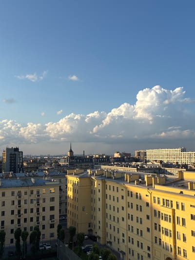 This is a high-angle, vertical-format photograph of a city skyline under a bright blue sky with large, fluffy white clouds on the left. The cityscape on the right is dominated by buildings with yellow facades, many featuring rows of windows and balconies. The sunlight casts long shadows, indicating it is likely late afternoon or early evening.
The scene captures an urban environment, possibly residential or commercial, given the scale of the buildings and their architectural details. The presence of trees and parked cars on the ground level suggests a developed area. A pointed spire or turret on one of the buildings is a notable architectural feature. The overall impression is one of a bustling city on a clear, sunny day. No people are clearly visible, and no text is discernible in the image. The location context suggests this is Annino, Russia.