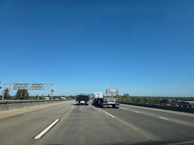 The image captures a bright, clear day on a multi-lane highway in West Sacramento, USA. The sky is a vivid, cloudless blue, indicating fair weather. The highway itself is paved and marked with white lines, with a concrete barrier on the left and a guardrail on the right separating the lanes from the surrounding environment.
Several vehicles are on the road. In the foreground, a black truck with a covered bed is in the left lane, followed by a white flatbed truck in the right lane. The flatbed truck has the license plate "4F29". Further ahead, other vehicles and trucks are visible, some disappearing into the distance. Overpasses and traffic signs are present, including a large overhead sign structure.
On the right side of the highway, beyond the guardrail, is a line of trees and some buildings. A billboard is visible in the distance, displaying the text "20 PLACES HONEST POTATOES". The overall scene suggests normal daytime traffic conditions. The perspective is from the driver's seat of a vehicle, looking forward along the highway.
