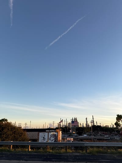 A wide shot presents a clear blue sky with faint contrails stretching diagonally. The horizon line showcases industrial structures, including towers and smokestacks, interspersed with vegetation and power lines. A billboard reading "WAG HOTELS" stands prominently. An American flag is visible. The image suggests an industrial area near a roadside.