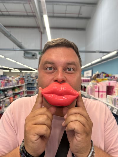 A man is holding a large, pink, novelty pair of lips over his own mouth in a retail environment. He is looking directly at the camera with his eyes wide open and a slight smile on his face. The man is wearing a pink t-shirt and several bracelets on his left wrist. The background consists of shelves stocked with various products, illuminated by fluorescent lighting, suggesting he is in a store, possibly a supermarket or a pharmacy. There is no indication that this image was taken in Palafrugell, Spain.