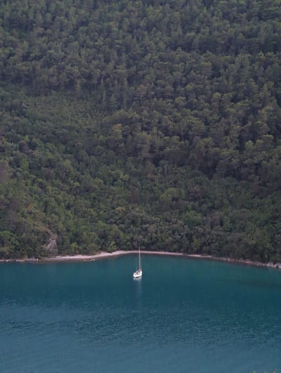 A sailboat with a white hull and a single mast is anchored in a tranquil, turquoise bay. The bay is nestled against a steep, densely forested hillside. The trees are primarily dark green, creating a lush, natural backdrop. The water appears calm with subtle ripples. A narrow strip of light-colored shore separates the water from the treeline. The scene suggests a secluded and peaceful coastal environment. The location is not within the city of Grad Labin, Croatia, but rather appears to be a natural bay.