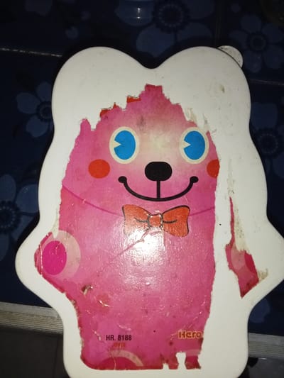 A flat, stylized, head-shaped decorative panel or toy is depicted in a top-down, slightly angled close-up. The panel primarily features a vibrant pink surface forming the face of a character, with large blue and black circular eyes, small red circular cheeks, a small black nose, and a black curved line forming a smile. An orange bow tie is positioned below the smile. This pink character is partially framed by a white, irregularly shaped outer layer, which shows extensive damage, peeling, and chipping along its edges, revealing the pink layer underneath. The visible surfaces of both the pink character and the white frame exhibit numerous scuffs, scratches, and dark smudges, indicative of wear. Faint, lighter pink circular patterns are present on the main pink body. In the lower left quadrant of the pink surface, text reads 'HR. 8188' and 'JOYCE' (partially obscured). To the right, 'Hero' is partially visible. The object rests on a dark blue surface adorned with a repeating pattern of lighter blue/grey floral or geometric motifs, consistent with a tiled floor or patterned fabric. A dark horizontal line is visible across the bottom of the background. The scene is located in Inxuba Yethemba Local Municipality, South Africa.