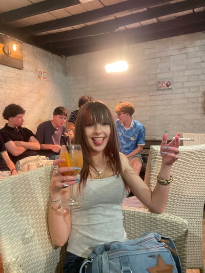 A young woman with long, dark hair and bangs is in the foreground of this medium shot, holding a glass of orange liquid and a cigarette. She is wearing a grey tank top, rings on her fingers, and several bracelets on her left wrist. Her tongue is sticking out, and she is looking at the camera. Behind her, three young men are seated, one in a black polo shirt, another in a dark purple t-shirt, and the third in a blue and white soccer jersey. The background consists of a white brick wall with two signs, wooden beams overhead, and wicker chairs. The overall setting appears to be an indoor bar or lounge. There is no specific information within the image to indicate the location in Hertsmere, United Kingdom.