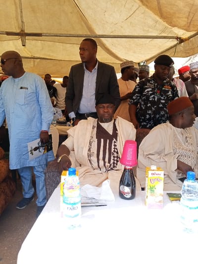 The image shows a group of men seated at a table under a white tent. The men are all dressed in traditional clothing, and some are wearing hats. There is a bottle of water, a bottle of juice, and a bottle of wine on the table, along with a pink plastic cup. Two of the men appear to be speaking to one another. The scene is taking place in the town of Zum, in the state of Borno, Nigeria, at the coordinates provided. The environment is dry and dusty with little vegetation. The tent is propped up on metal poles, with the white fabric stretched taut.