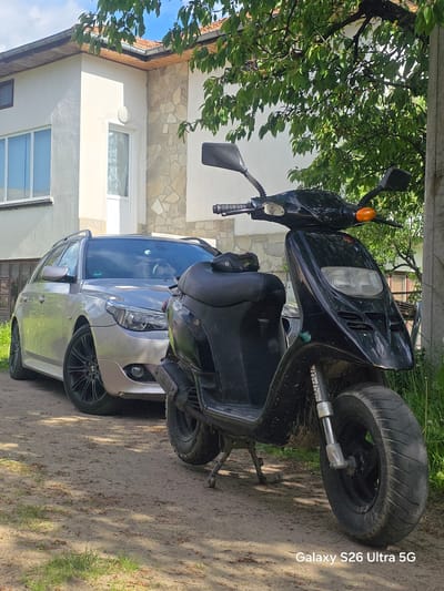 The image depicts a black scooter parked in front of a silver BMW station wagon. The vehicles are located on a dirt area with some grass. A residential building with a white facade and visible windows stands in the background. A tree with green leaves is partially visible above the scooter. The photo appears to have been taken outdoors, possibly in a residential or suburban area in Barutin, Bulgaria. There are no people visible in the frame.