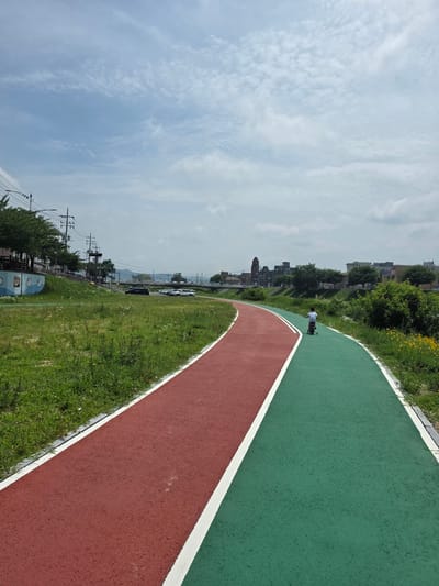 A young boy rides a tricycle on a paved bike path that is divided into a red lane and a green lane. The path curves to the right and runs alongside a grassy field. In the distance, there are buildings and trees, and a bridge crosses a body of water. The sky is cloudy. The image appears to be taken in Anseong-si, South Korea, possibly in a park or recreational area along a river.