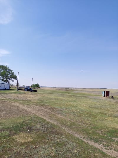 The image depicts a rural landscape under a clear blue sky with a few wispy clouds. The scene is set in a wide, grassy field in Akzhar, Kazakhstan.

In the foreground, on the left side of the image, is a white building with a light-colored roof and a large green tree. Next to the building, a blue tractor is parked. Utility poles with wires run across the field. The ground is covered with dry, matted grass and some patches of greener vegetation. Tire tracks are visible in the dirt.

In the middle ground, the field stretches out, appearing mostly flat with some gentle undulations. A small, rustic wooden structure with an open doorway and a rusty metal roof stands in the right portion of the frame. A wheelbarrow is positioned near this structure.

The background shows a distant horizon with a few low hills and scattered trees. The overall impression is one of a serene, open countryside. The time of day appears to be late morning or early afternoon, judging by the bright sunlight and the long shadows. The weather is clear and likely warm, suitable for outdoor work in an agricultural setting. There is no visible text in the image.