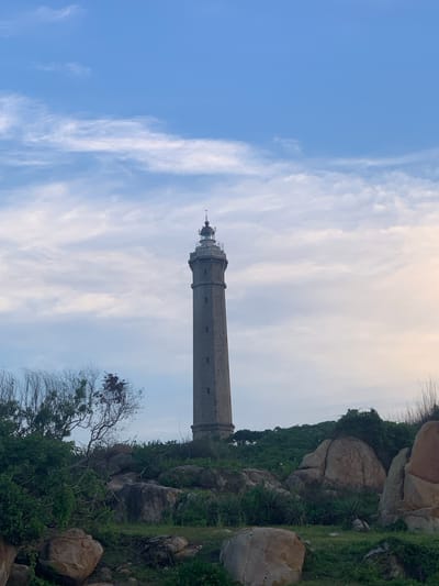 A lighthouse stands tall against a backdrop of a partly cloudy sky. The lighthouse is a cylindrical stone structure with a metallic lantern room at the top. It is situated on a rocky, green hillside. The terrain around the lighthouse is uneven, with large boulders and patches of vegetation. The sky displays a mix of blue sky and soft, wispy clouds, suggesting it might be late afternoon or early morning. The overall scene evokes a sense of isolation and natural beauty, characteristic of coastal or elevated locations. There is no visible text in the image.