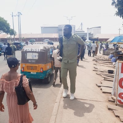 The image depicts a street scene in Kufang, Nigeria (latitude: 9.863, longitude: 8.867). A man in a green uniform walks towards the right of the frame. A woman in a pink dress and black handbag walks towards the left of the frame. The woman's back is facing the viewer. A yellow and brown three-wheeled auto-rickshaw is parked on the left side of the frame. The auto-rickshaw is labeled "PTA-7647" and "HASKE." A man in a white t-shirt and blue jeans stands next to the auto-rickshaw, facing the viewer. There is a stack of wooden planks in the lower right of the image. Other people can be seen in the background of the image. The street is paved and there are power lines overhead. The sky is clear and blue. The environment appears to be a busy street scene in a small town or village. The scene suggests that it is a hot, sunny day. The time of day is likely late morning or early afternoon.