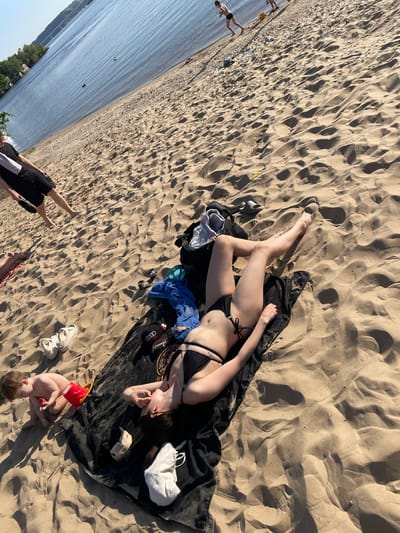 A woman in a black bikini lies on a dark-colored blanket on a sandy beach. A young child in a red swimming suit sits near the blanket, playing with a red bucket. Other items scattered around the blanket include a white bag, a white baseball cap, shoes, a blue towel, and a black hat. In the background, people are visible in the water. The location is Матюшино, Russia, based on the landscape.