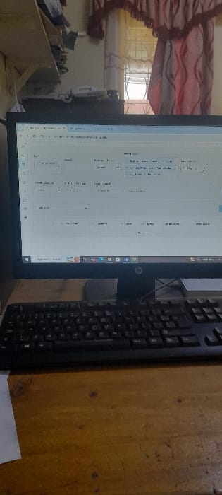 The image shows a desktop computer with a black keyboard in front of it. The computer is displaying a spreadsheet program with data and various fields. The desktop is wooden and a piece of white paper is visible in the bottom left corner.  The room is simple, featuring a window with a curtain behind the computer. The location is a desk in a small office in Njoro, Kenya, based on the provided coordinates.