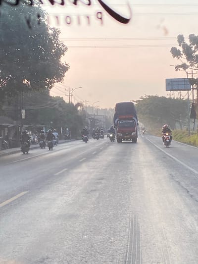 The image depicts a street scene in Jekulo, Indonesia, likely during the daytime given the ambient light. The foreground is dominated by a paved road with visible tire tracks and yellow lane markings. A convoy of motorcycles, some with riders wearing helmets, are traveling on the road.
In the middle ground, a red truck carrying a dark-colored, oversized load is moving forward. More motorcycles are visible in the distance, forming a procession. The left side of the road features a line of buildings and trees. The right side of the road is bordered by trees and a sidewalk, with utility poles and wires extending into the distance. A billboard-like structure is also present on the right. The background shows a hazy sky, suggesting either early morning or late afternoon light, or possibly atmospheric conditions. The overall impression is of a busy thoroughfare with significant motorcycle traffic.