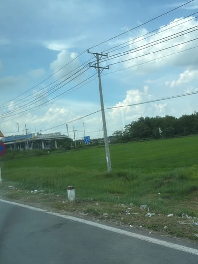 The image depicts a roadside scene in Long Thành, Vietnam, with a focus on a toll booth complex visible in the background. A bright green, cultivated field, likely rice paddies, stretches across the midground. A tall, utility pole with multiple wires runs diagonally through the frame, dominating the sky. The sky itself is partly cloudy with patches of blue. A paved road occupies the foreground, with a white line marking the edge. Along the roadside, there are scattered pieces of white litter. A white and red marker post stands on the grassy verge. The toll booth area features multiple lanes, signage, and overhead structures. Trees line the horizon behind the field. The overall impression is of a rural highway environment in Vietnam.