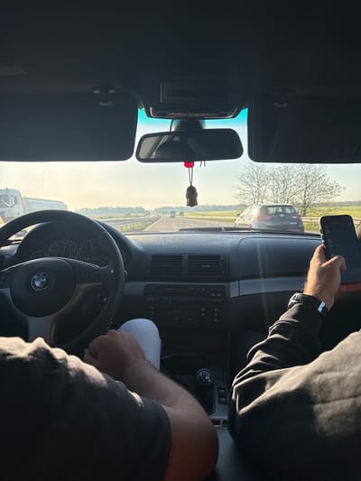 The image depicts the interior of a BMW car, likely in motion, on a highway near Opalchenets, Bulgaria, suggested by the road signs visible through the windshield. The driver is holding a mobile phone in one hand while gripping the steering wheel with the other. A passenger is partially visible on the right side of the frame. The dashboard and central console features, including the air conditioning vents and radio, are clearly displayed. The rearview mirror and a decorative wooden ornament hanging from it are visible in the upper left. Outside the car, other vehicles and open rural landscapes are shown under a slightly hazy daylight sky.