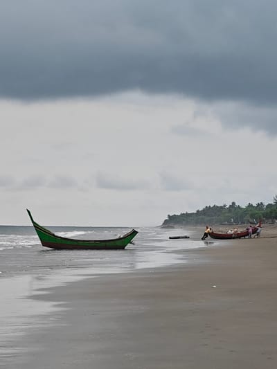 The image depicts a beach scene with overcast skies. In the foreground, a green and red fishing boat rests in shallow water near the shore. Further down the beach, several people are gathered around another red fishing boat, appearing to push or pull it onto the sand. The background shows a line of lush green trees and a rocky outcrop. The wet sand reflects the cloudy sky. The scene is likely the coast of Uleekareung, Indonesia, a region known for its beaches and fishing communities.