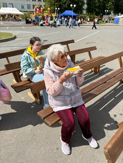 A high-angle shot shows two individuals seated on a wooden bench. One, possibly an elderly person, wears an old-age makeup prosthetic and costume, with a white wig, large sunglasses, and a white puffy vest over a pink sweater, maroon pants, and white sneakers. The second person, likely a younger individual, is seated to the left of the first. They are wearing eyeglasses, a yellow shirt, and a light blue jacket. Both individuals are holding food, which appears to be corn on the cob, in their hands.

The wooden bench consists of multiple planks of wood arranged at varying heights. The scene is situated in an open, paved area, likely outdoors, with other structures and people visible in the background. In the distance, there is a playground with an inflatable structure, several tents, and a number of people walking around. A multi-story building is visible on the left-hand side of the frame.

Based on the available information, the location is identified as Valozhyn, Belarus.