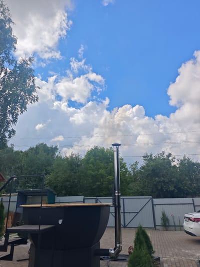 The image depicts an outdoor scene under a bright blue sky with scattered white clouds, likely in the SNT Luzhniki area of Russia. In the foreground, a large black, barrel-shaped object, possibly a hot tub or a large grill, is partially visible, with a wooden rim and a metal chimney pipe extending upwards. Adjacent to it is a metal staircase. Behind the object, a grey metal fence stretches across the scene, separating the yard from a paved area where a white car is parked. Several small evergreen trees are planted in front of the fence. The background is filled with lush green trees, suggesting a semi-rural or suburban setting. Power lines are visible horizontally across the middle ground, intersecting with the trees.