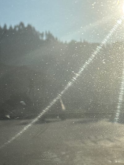 The image depicts a hazy, sunlit scene, possibly taken through a window or a dirty surface, with numerous small specks of light scattered across it. In the background, a blurred silhouette of trees against a bright sky is visible. A diagonal streak of light, possibly caused by the sun's glare or a physical scratch on the surface, cuts across the image. The foreground appears to be a darker, textured surface, possibly asphalt or pavement. A faint, blurred human figure is visible in the middle ground, appearing to be in a seated or crouched position. The overall impression is one of a somewhat degraded or obscured view of an outdoor environment, lacking specific identifying landmarks of Smilyan, Bulgaria.