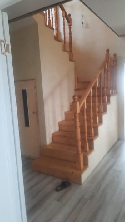 A wooden staircase is visible in a residential interior, likely in the city of Қаражар, Kazakhstan. The staircase, constructed from light-colored wood, features carved balusters and a wooden handrail. The walls surrounding the staircase are painted a neutral, light beige color. To the left of the staircase, a door with a narrow, dark rectangular window is present. The floor is covered with light gray laminate flooring with a wood grain pattern. A single dark slipper is positioned on the floor near the bottom of the staircase. The lighting suggests it is daytime, with natural light entering from an unseen source, casting shadows on the stairs and floor.