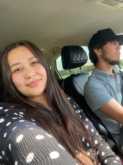 Two people are in a car. In the foreground, a woman with long, dark brown hair and a black and white polka dot shirt is smiling at the camera. Behind her, a man with dark hair and a beard, wearing a black cap and a gray collared shirt, is looking out the window. The car appears to be moving, as indicated by the slight blur in the background. The environment outside the car window suggests a rural or suburban setting with trees and a cloudy sky. The interior of the car shows black leather seats. The image was taken inside a vehicle, likely in Gornyi, Russia, given the provided context.