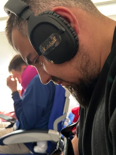 A bearded man wearing black Marshall headphones is shown from the side in an airplane seat. Another passenger is visible in the background. The image suggests the interior of a commercial aircraft, with elements like a headrest and blue seat fabric visible. There is no indication of the specific location of Stolbishche, Russia in the image.