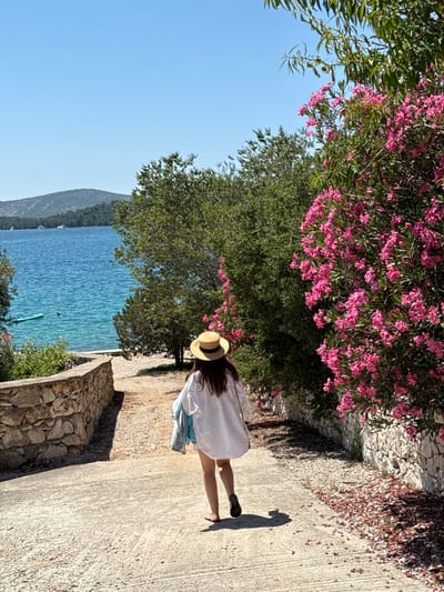 A person wearing a white oversized shirt, shorts, a straw hat, and dark sandals walks down a paved pathway towards the sea in Grad Šibenik, Croatia. The person is carrying a light blue and patterned fabric over their left arm. The pathway is lined with a low stone wall on the left and vibrant pink oleander bushes on the right. Beyond the pathway, a tranquil blue sea is visible, with a green paddleboard and a swimmer in the water. In the distance, lush green hills and islands under a clear blue sky form the backdrop. The scene suggests a warm, sunny day.