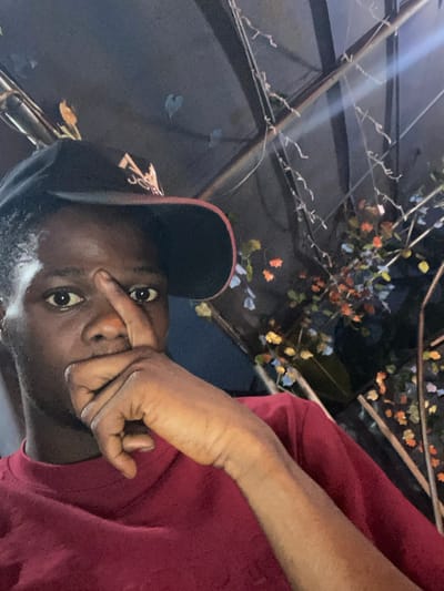 The image depicts a solitary individual, identified as male, with dark skin, captured in a selfie-style composition. He is wearing a black baseball cap featuring a red underside brim and a light-colored emblem on the crown. A maroon t-shirt covers his upper torso. His right hand is positioned with the index finger touching the bridge of his nose, while the remaining fingers obscure his mouth. His eyes are directed straight ahead at the camera. The background presents a dimly illuminated environment under what appears to be a covered structure. Visible elements include dark fabric draping over metal framework, string-like decorations, and artificial leafy branches exhibiting orange, yellow, and blue hues. A bright light source is reflected in the upper right portion of the frame. The image contains no intrinsic visual information to confirm a specific geographical location within Igbessa, Nigeria, or elsewhere.