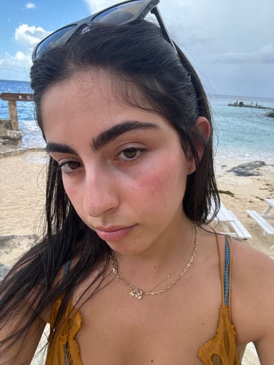 A close-up portrait shows a female individual in a tropical beach environment, specifically at Buccanos Beach Club in Cozumel, Mexico. The subject, positioned centrally, has dark brown hair, dark brown eyes, and prominent eyebrows. She is wearing a golden-brown or mustard-colored lace-trimmed top with blue straps, accessorized with two gold necklaces; one is a thicker chain with a cluster of small flower-like charms, and the other is a thinner chain with a single small charm. Dark sunglasses are perched on top of her head. Her gaze is directed towards the camera.
In the background, to the left, a weathered wooden sign partially displays the white letters "B U" on a plank, consistent with "Buccanos." Behind this sign, light blue-green ocean water is visible. To the right, beyond the subject's shoulder, a sandy beach area includes at least two white wooden picnic tables or benches. Further out in the water, a distant rocky outcrop or small platform is discernible. The sky above is partly cloudy with patches of blue. The immediate foreground consists of sandy terrain.