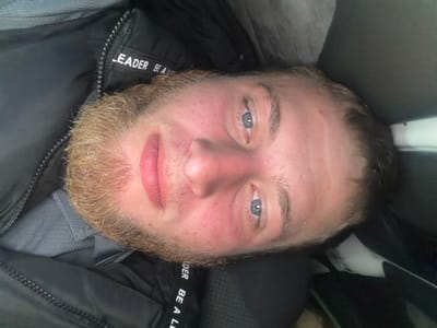 A close-up, rotated selfie of a young Caucasian male with light brown hair and a light brown beard is shown. He has blue eyes, fair skin with some redness, and pink lips. He is wearing a dark jacket with a zipper and the words "LEADER BE A L" visible on a white label, and a grey collared shirt underneath. The background appears to be dark and possibly indoors, with a greyish area on the upper left and a black area on the right. The image is oriented such that the man's head is towards the bottom of the frame. There is no discernible indication of the city of Sculeni, Moldova within the image itself.