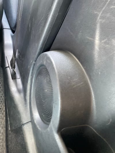 The image shows a close-up view of the interior of a vehicle, specifically focusing on the lower section of a door panel. A black speaker grille is visible, recessed into the plastic paneling. To the left of the speaker, a black strap with a metallic clasp is attached to a rectangular slot in the panel. The surrounding plastic appears to be dark gray and shows signs of wear, with light scuff marks visible. The lighting suggests an indoor environment or a shaded outdoor location. The background is dark and out of focus, but hints at other parts of the car's interior, possibly carpeting. There are no people or discernible action in the image. The scene is devoid of any specific urban context from Tyulyugul, Russia.