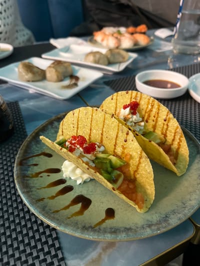 The image displays an elegantly presented meal served indoors, likely in a restaurant in Basiglio, Italy. The primary focus is on two hard-shell tacos artfully arranged on a round, light grey-green ceramic plate with a distinctive patterned rim.

Each taco is filled with a colorful array of ingredients: diced reddish pieces (possibly salmon or tomato), cubed avocado, dollops of white creamy sauce (such as sour cream), and a bright garnish of small red spheres, resembling fish roe or caviar. A dark, glossy sauce is drizzled decoratively across the tacos and onto the plate, adding to the visual appeal.

In the softly blurred background, additional dishes suggest a varied dining experience. To the left, a long, narrow white dish holds several prepared shrimp or prawns. Below it, two other rectangular white dishes contain multiple round, brownish items, which appear to be dumplings or gyoza. A small white oval bowl holds a dark dipping sauce, possibly soy sauce. The table is set with a dark, textured woven placemat over a blue surface. The lighting is soft, indicative of an indoor dining atmosphere. There is no visible text or specific time of day discernible from the image.