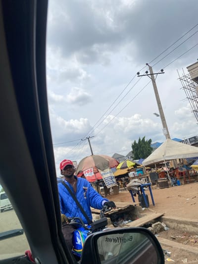 This image depicts a bustling roadside market area in Naze, Nigeria, under an overcast sky with numerous light grey clouds. The perspective is from inside a vehicle, with its dark frame visible along the left edge and a rearview mirror showing the reflection of "Be different" in the lower right foreground.
In the mid-ground, a male individual is prominently featured, seen from approximately the waist up. He wears a red baseball cap with "Yankees" in white script, a blue quilted jacket unzipped to reveal a dark garment, and black fingerless gloves. A dark strap crosses his chest. He has short dark hair and facial hair, and is looking directly towards the camera. He is positioned on or next to a blue and white Honda motorcycle, partially visible beneath him, with his hands near the handlebars.
The background shows a street lined with multiple vendor stalls, characterized by various colored umbrellas providing shade, including red, orange, brown, yellow, and white. One prominent sign on a red/brown umbrella reads "POS HERE," with another sign below it listing "SERVICES" such as "Withdrawal, transaction, copy Bills, buy Airtime, Recharge Card." Further back on the right, a large white tent or canopy covers several wooden tables and stools, indicating a more extensive selling area. Utility poles with power lines crisscross the upper portion of the scene, and a streetlight is affixed to one pole. To the far right, a building under construction with visible scaffolding is present. The ground appears uneven and unpaved in sections, with some debris visible near the stalls. A cluster of green trees stands in the mid-distance behind the market activity.