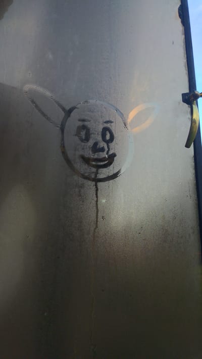 This image captures a stylized face drawn onto a condensation-covered transparent surface, identified as a window, within the City of Longuo 'A', Tanzania. The drawing, created by wiping away moisture, features a circular head with two prominent, elongated ears extending laterally. Facial elements include two oval eyes with small pupils, a triangular nose, and an upward-curving mouth. A thin stream of liquid originates from the lower center of the mouth and descends vertically across the lower half of the drawing. The entire surface is heavily misted with numerous small water droplets. On the far right, a dark vertical frame element is visible, to which a golden-colored, curved handle is affixed. The ambient lighting appears diffused through the condensation. No human figures are present, only the static depiction on the glass.
