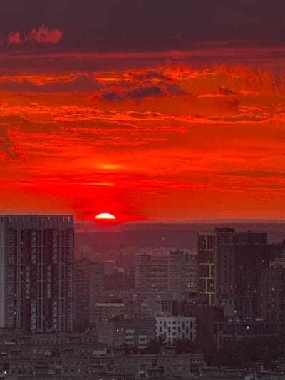 The image shows a vibrant, fiery sunset over the city of Savenki, Russia. The sky is dominated by intense shades of red and orange, with streaks of dark purple at the top. Wispy clouds are illuminated by the setting sun, creating a dramatic and atmospheric effect.

In the foreground, the silhouette of a city skyline is visible. Several buildings, including a tall, modern high-rise on the left and a cluster of shorter, more densely packed buildings on the right, are silhouetted against the bright sky. The details of the buildings are somewhat obscured by the low light and the atmospheric conditions.

The sun itself is a bright, red disc, partially obscured by the horizon, casting a warm glow over the entire scene. The overall impression is one of a powerful natural spectacle occurring above a bustling urban landscape. There are no people or animals visible in the image.