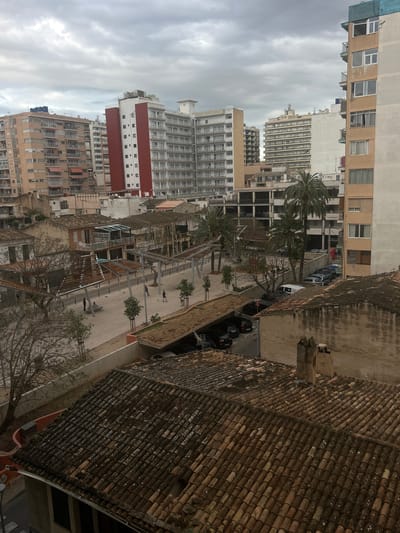 The image shows an elevated view over part of S'arenal, Spain. The foreground contains tiled roofs. Beyond that is a promenade with trees and scattered people. On the left, there are buildings of varying heights, including apartment blocks. Parked cars are visible. The sky is overcast.