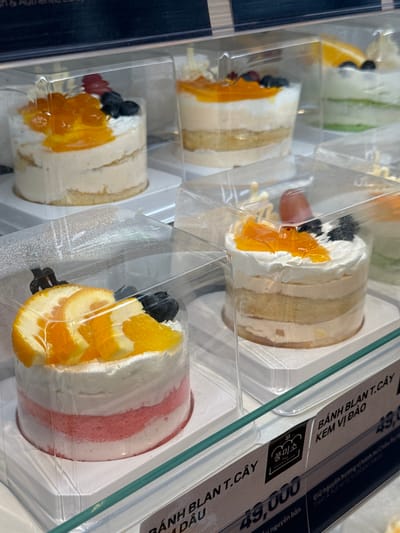The image displays a close-up view of several individual cakes presented in clear plastic packaging, arranged on shelves within a retail display case. The cakes are tiered, with visible layers of sponge cake and cream. Some cakes are topped with fruit such as sliced oranges and berries, while others feature a glossy fruit topping. The packaging is consistent across all visible cakes, suggesting they are part of a product line.
Below the display case, a price tag is visible, indicating "BÁNH BLAN T. CÂY KEM VỊ ĐÀO 49,000". This translates to "Cream Peach Flavor Cake 49,000" with a price of 49,000. Another price tag is partially visible, showing "49,000" and text that includes "bánh nguyên bản". A logo for "FOONG MISO" is also present on a sticker. The text "Fresh & Authentic European bakery" is visible on another sticker.
The lighting suggests an indoor commercial environment, likely a bakery or a patisserie. The overall scene is clean and organized, emphasizing the presentation of the desserts. There are no people visible in the frame. The image appears to be taken in Phu Thuy, Vietnam, as indicated by the Vietnamese text on the price tags.