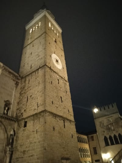 An upward angle depicts a tall, stone clock tower at night. A clock face is visible midway up the tower, illuminated from below along with the tower's stone surface. The upper portion features lighted windows. To the right, another building with crenellations is partially visible, also lit from below. The background is a dark night sky.