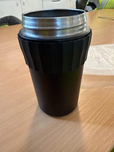 A close-up, eye-level shot shows a black, cylindrical object with a silver rim on a wooden table. The object is roughly in the center of the frame and occupies the majority of the image. The silver rim is textured with two distinct horizontal ridges. Below the rim, the black body of the object has a ribbed, segmented pattern that tapers downwards. The object appears to be hollow. The wooden table surface has a light, grainy texture with visible wood grain lines. In the background, a white, crinkled plastic bag is visible to the right of the object. To the left of the object, out of focus, a dark surface with a hint of yellow and black fabric is visible, potentially clothing or safety gear. The overall lighting suggests an indoor environment. This scene is located in the city of Södra Svartbyn, Sweden.