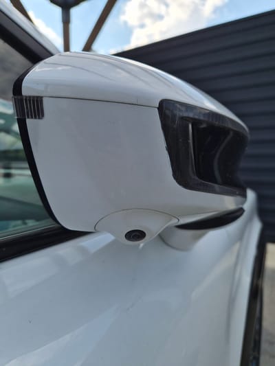 A close-up, low-angle, shallow depth-of-field shot shows a white car's side-view mirror. A small camera lens is visible on the bottom of the mirror housing. The mirror housing is white and has a black accent around the reflective surface. The background includes a dark corrugated wall, some wooden beams, and a blue sky with white clouds. The image is taken in Lenina, Russia.