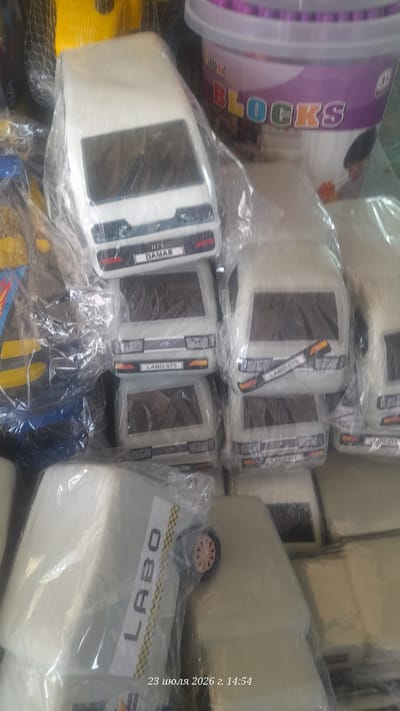 This is a close-up, eye-level, indoor photograph of several toy vehicles, likely a fleet of small, white vans, neatly arranged and individually wrapped in clear plastic. The toys are the primary focus, dominating the frame. Some of the toy vehicles have labels on their front grills. One label reads "0/1 DAMAS," and another reads "LABO 075." The toys are stacked or placed side-by-side, suggesting they are either for sale or storage. In the background, partially visible, is a large, purple and white plastic tub with the word "BLOCKS" printed on it in colorful letters, suggesting it might contain toy building blocks. The lighting is somewhat dim, and the overall impression is that of a commercial setting, possibly a shop or warehouse in Shurchi, Uzbekistan. The timestamp "23 июля 2026 г. 14:54" is superimposed on the bottom right of the image, indicating the date and time the photograph was taken.
