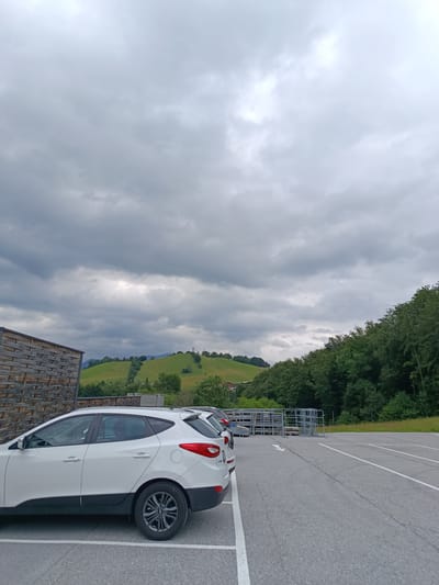 The image depicts an outdoor parking lot with several white cars parked. In the background, there is a lush green hill dotted with trees, and a cloudy sky overhead. To the left, a building with a wooden facade is visible. In the far distance, mountains can be seen. The parking lot is paved with asphalt and marked with white lines. A metal structure, possibly for storage or a fence, is present in the middle ground. The overall scene suggests a location in Hallein, Austria, given the characteristic mountainous terrain and the architectural style of the wooden building.