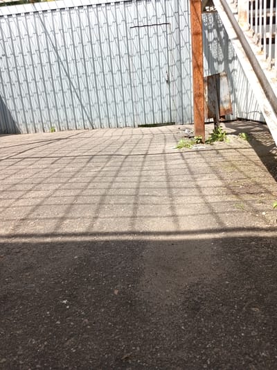 This image depicts an outdoor scene with a corrugated metal fence in the background. A rusty metal pole stands in front of the fence, with a small, detached metal piece leaning against it. The ground is paved with asphalt, and a grid-like shadow pattern is cast across it, likely from a fence or grating. Some sparse green vegetation is growing near the base of the metal pole. The scene appears to be in an industrial or utilitarian area, possibly within the city of Furmanov, Russia, given the context provided. There are no people or animals visible in the image.