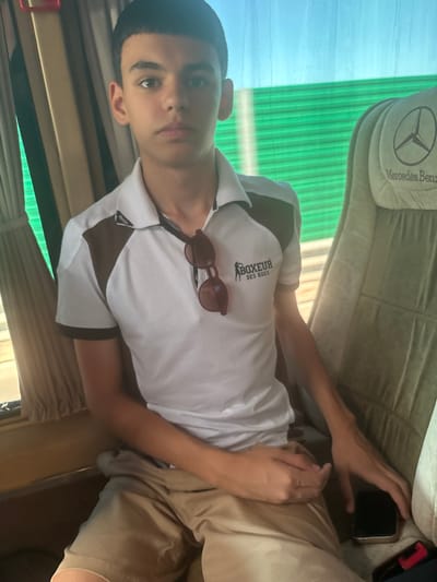 A young male adolescent is seated in the passenger seat of a Mercedes-Benz bus, looking directly at the camera. He is wearing a white polo shirt with brown accents on the shoulders and sleeves, and tan shorts. A pair of sunglasses is casually placed on the collar of his shirt. His right hand rests on his lap, near a smartphone placed on the seat next to him. The bus interior is visible, with beige curtains on the left and the distinctive green, horizontally slatted blinds of the window on the right. The headrest of the seat behind him bears the Mercedes-Benz logo. The lighting suggests it is daytime. The location of Fushë-Krujë, Albania is not discernible from the visual elements in the image.