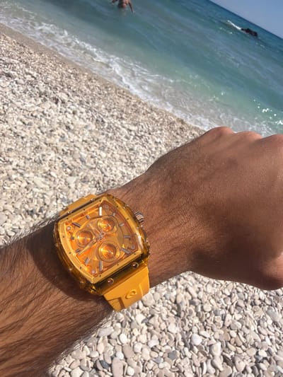 A person's arm, wearing a vibrant orange digital watch, rests on a pebble beach. The sea, with gentle waves and a rocky outcrop in the distance, is visible behind the beach. The scene is set on a bright, sunny day.