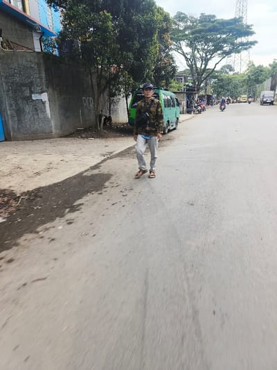 A man walks toward the camera on a paved road. He wears a camouflage jacket, grey sweatpants, sandals, and a black cap. A black shoulder bag is slung across his chest. Behind him, a green van is parked alongside the road. Further down the street, motorcycles and a truck are visible. Trees line the street, and a communication tower rises in the background. A grey wall borders one side of the road.