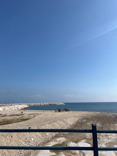 The image shows a beach in Limenaria, Greece. The location is a small bay near the harbor, identified by the coordinates 40.626 latitude and 24.573 longitude. The foreground is a sandy beach with a large concrete structure, or jetty, in the middle. There are white rocks on the structure. The background is a blue ocean. The horizon is marked by a small strip of white sand. The sky is clear and blue. A group of people are gathered on the beach, interacting with each other, near the jetty. There is a large black metal railing in the foreground that is partially obscuring the view of the beach. The sand on the beach is light brown and rough, with some rocks and debris visible. The image appears to have been taken during the day.